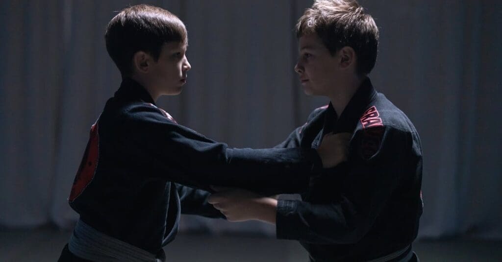 Two boys in black martial arts uniforms practicing teen self-defense skills by working on upright grappling and shirt-grab counters.